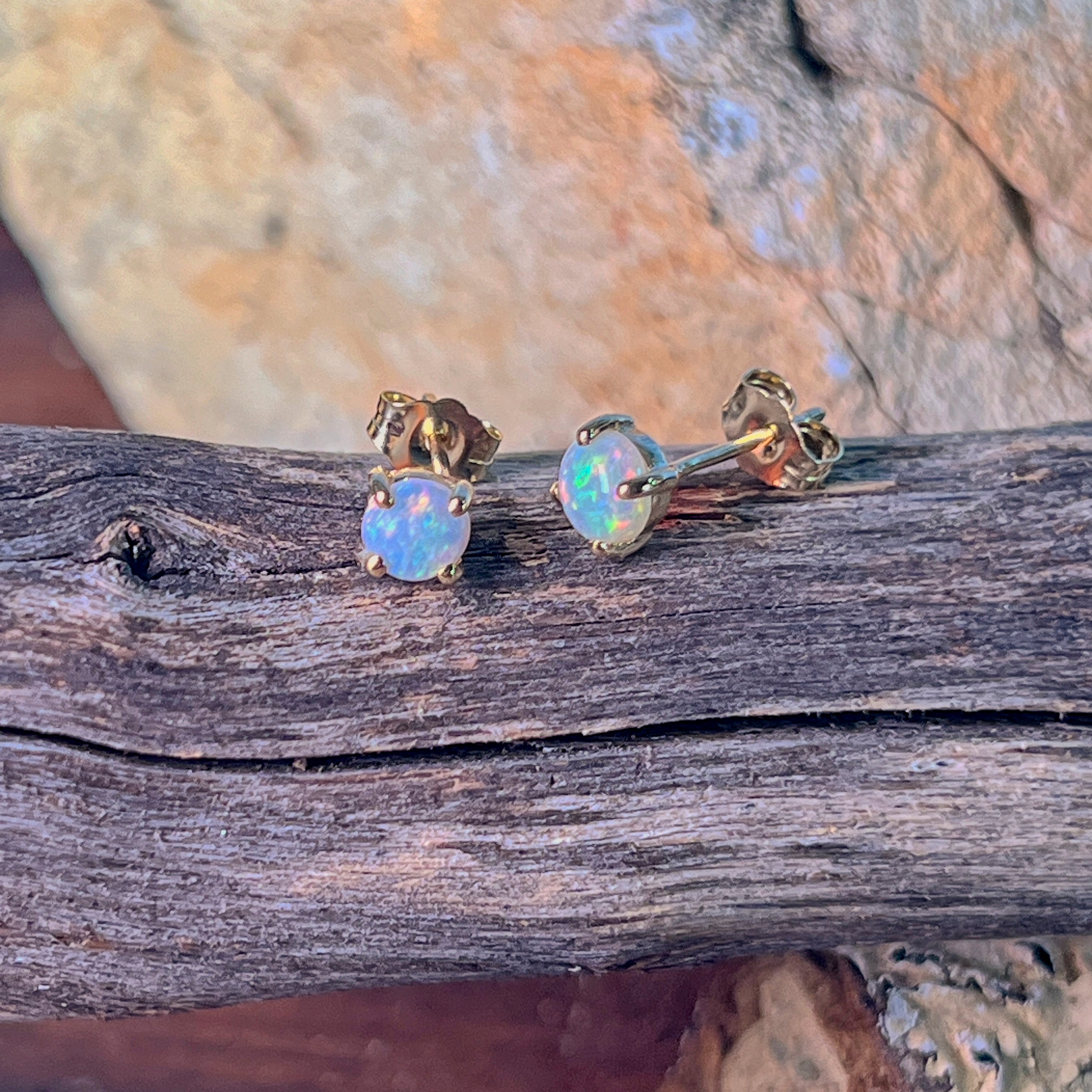 Round crystal opal stud earrings in 9kt yellow gold resting on driftwood